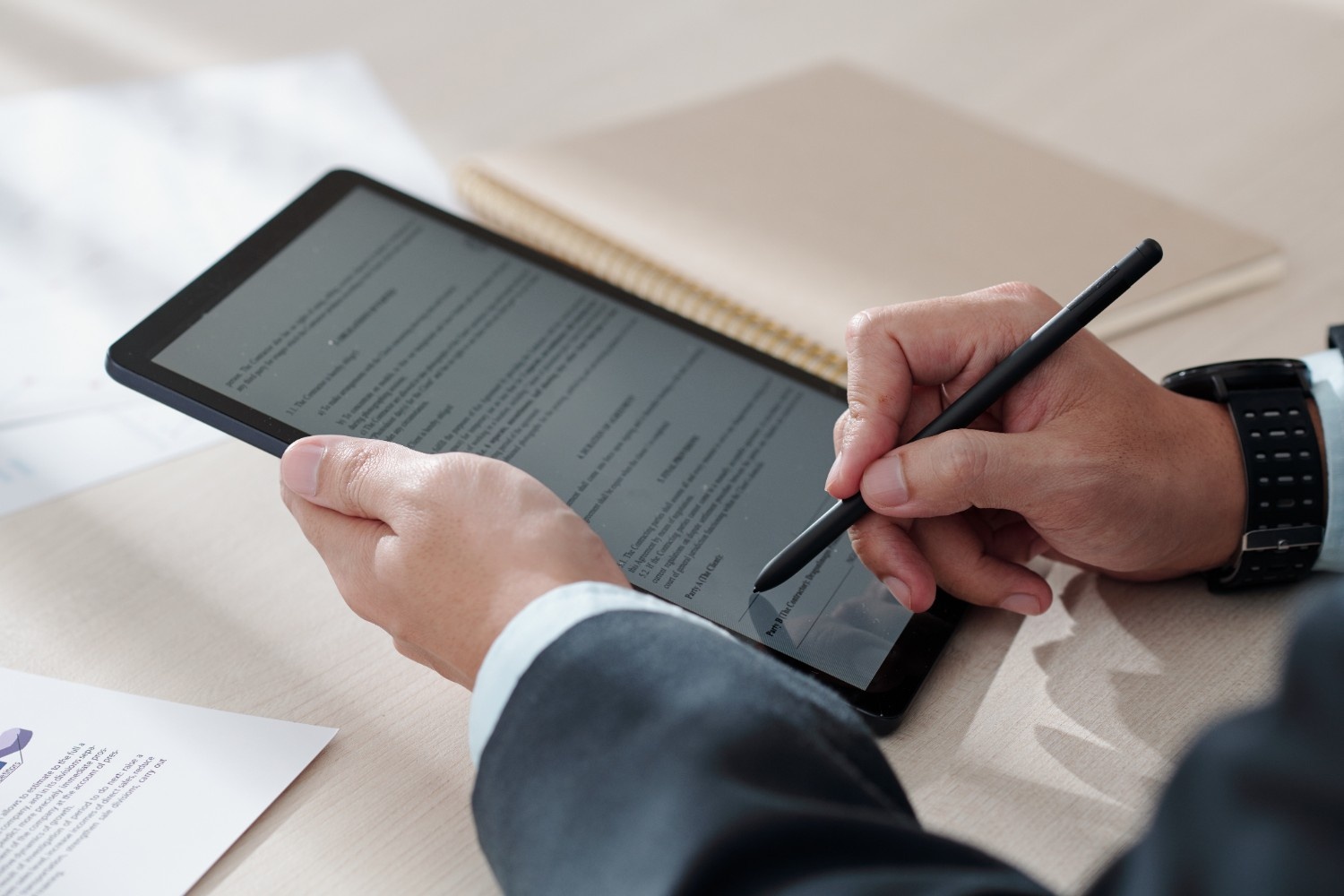Business professional using stylus pen on digital tablet displaying document text, with paper documents and notebook visible on desk, wearing watch and dark suit