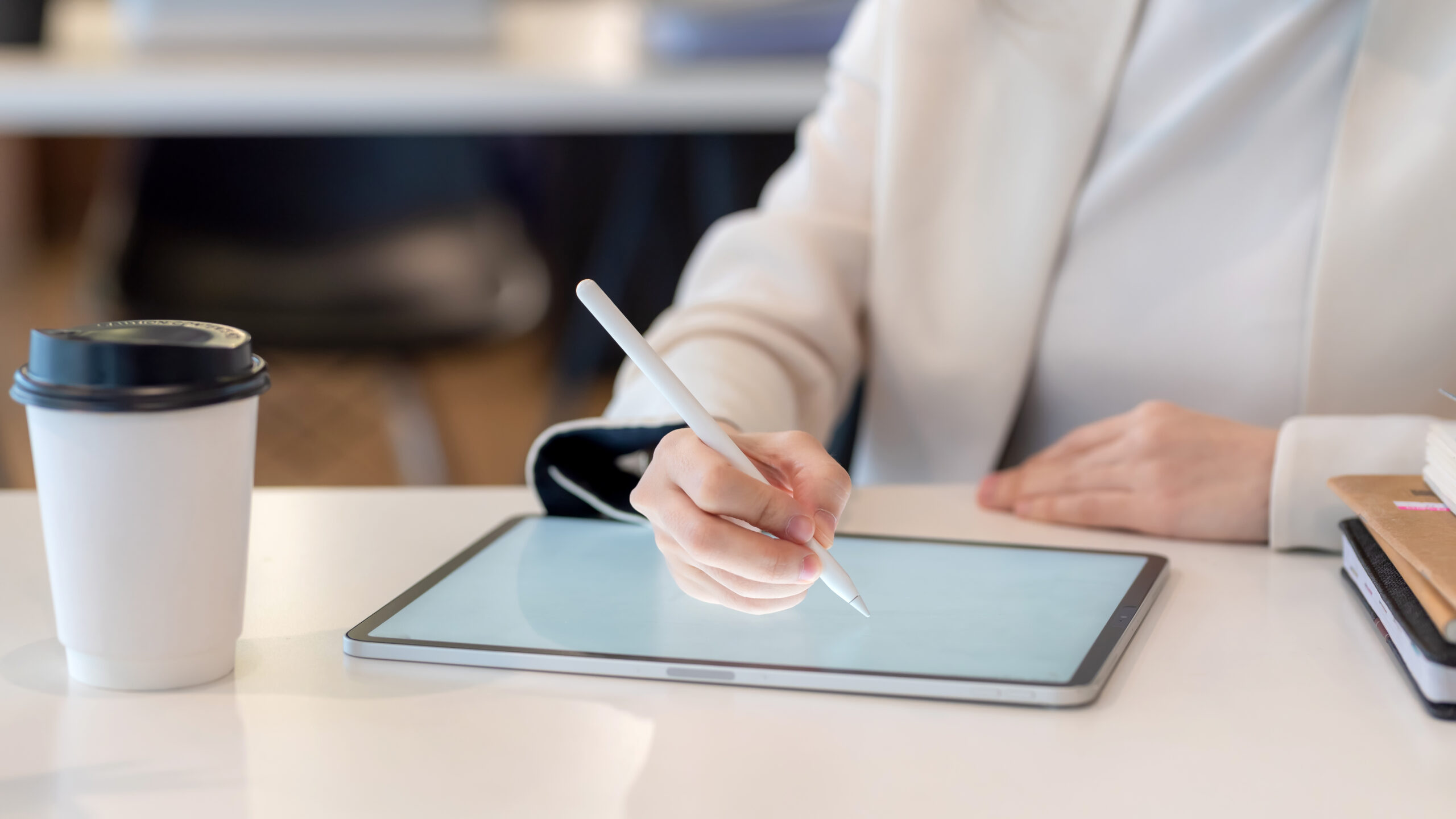 Professional in white blazer using stylus on digital tablet for eSignatures, with coffee cup, smartphone, and notebook in a modern office.