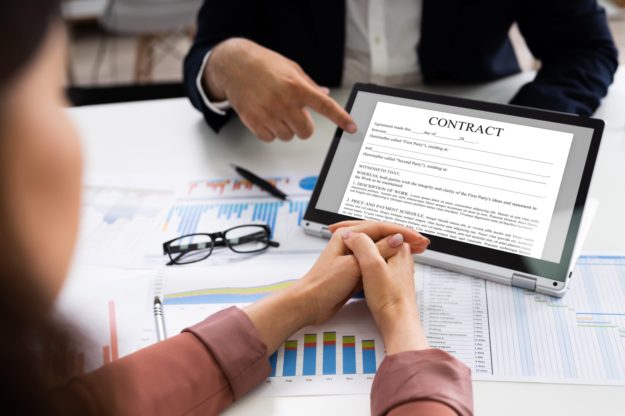 Business professionals reviewing a contract document displayed on a tablet screen during a meeting, with financial charts, graphs, and eyeglasses on the desk, one person pointing to the contract while another listens