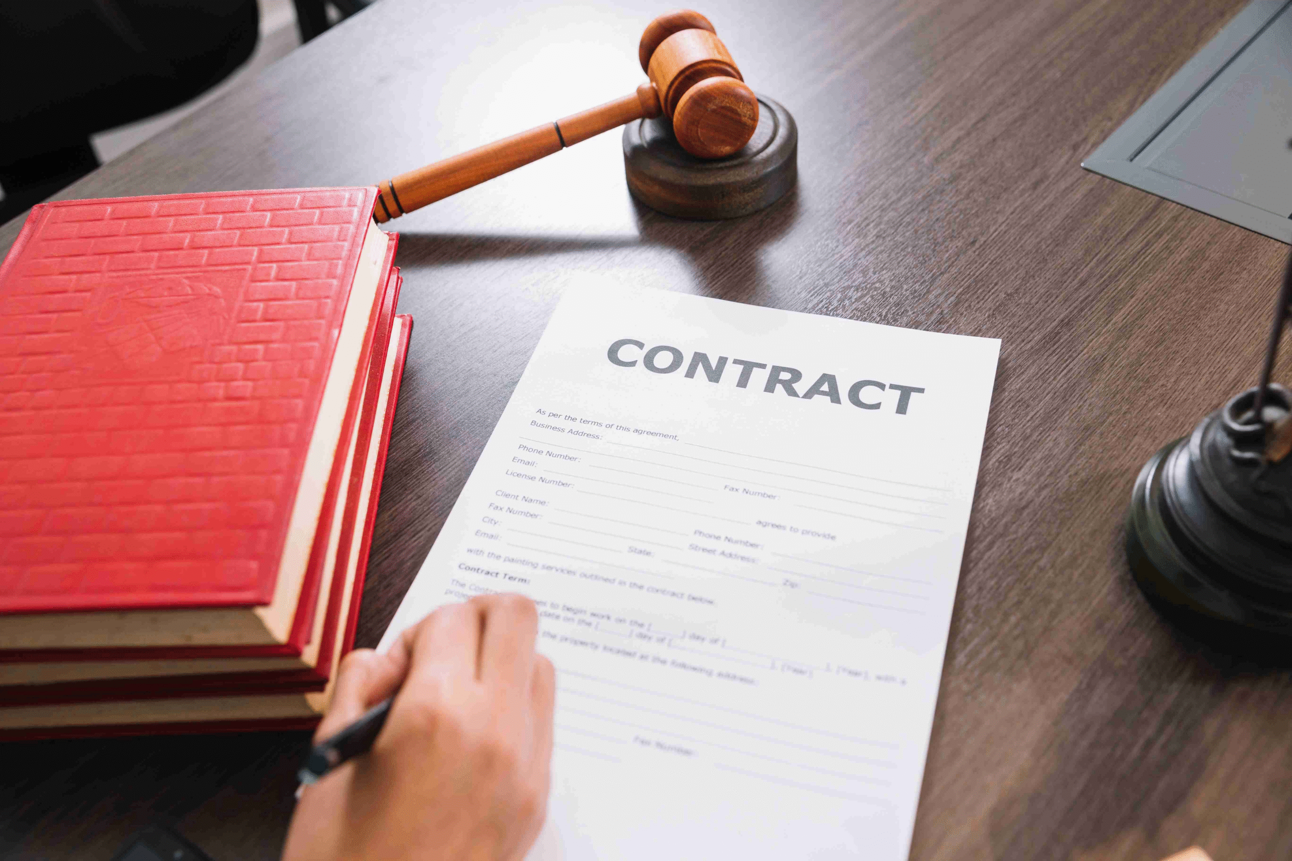 Person signing legal contract document on desk with wooden gavel, red law books, and laptop in background