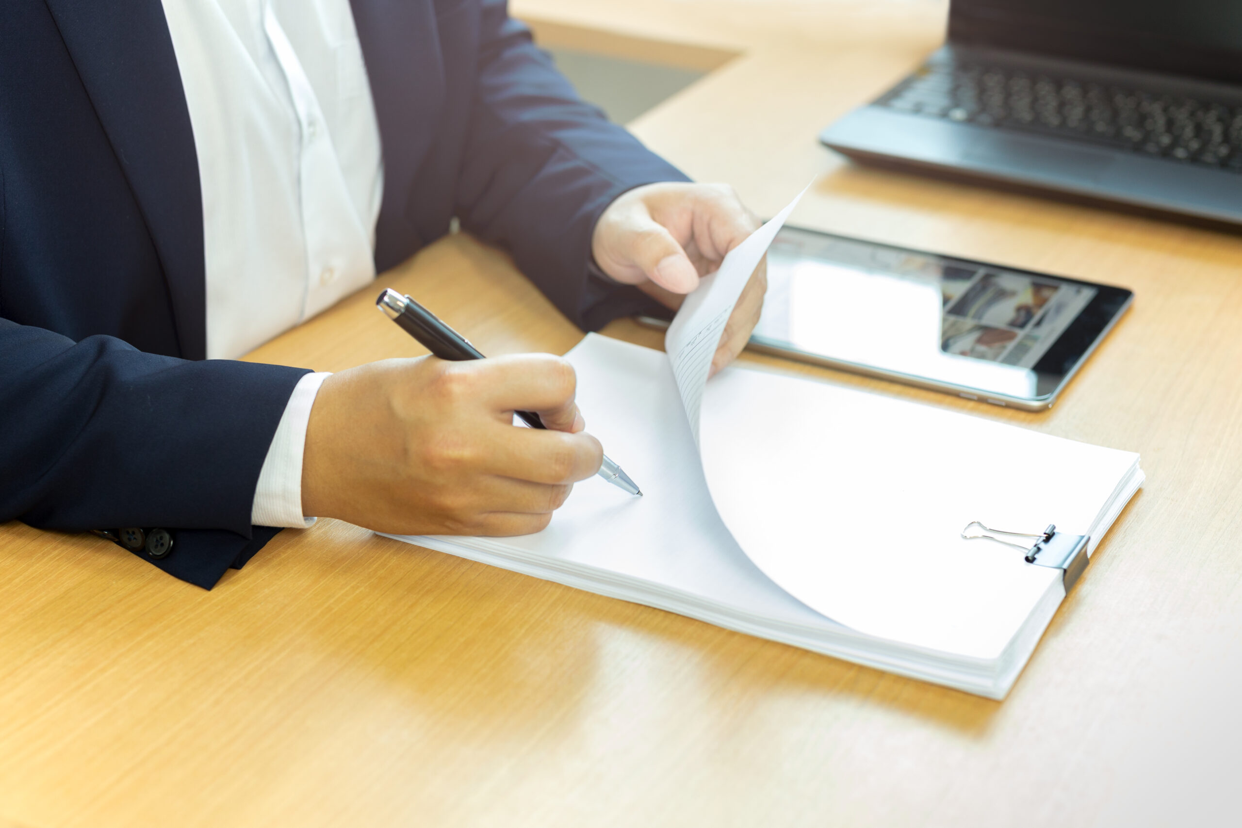 Two business professionals in suits reviewing documents together at a wooden desk, with one person writing on papers while the other points to a document, a tablet and laptop visible in the background