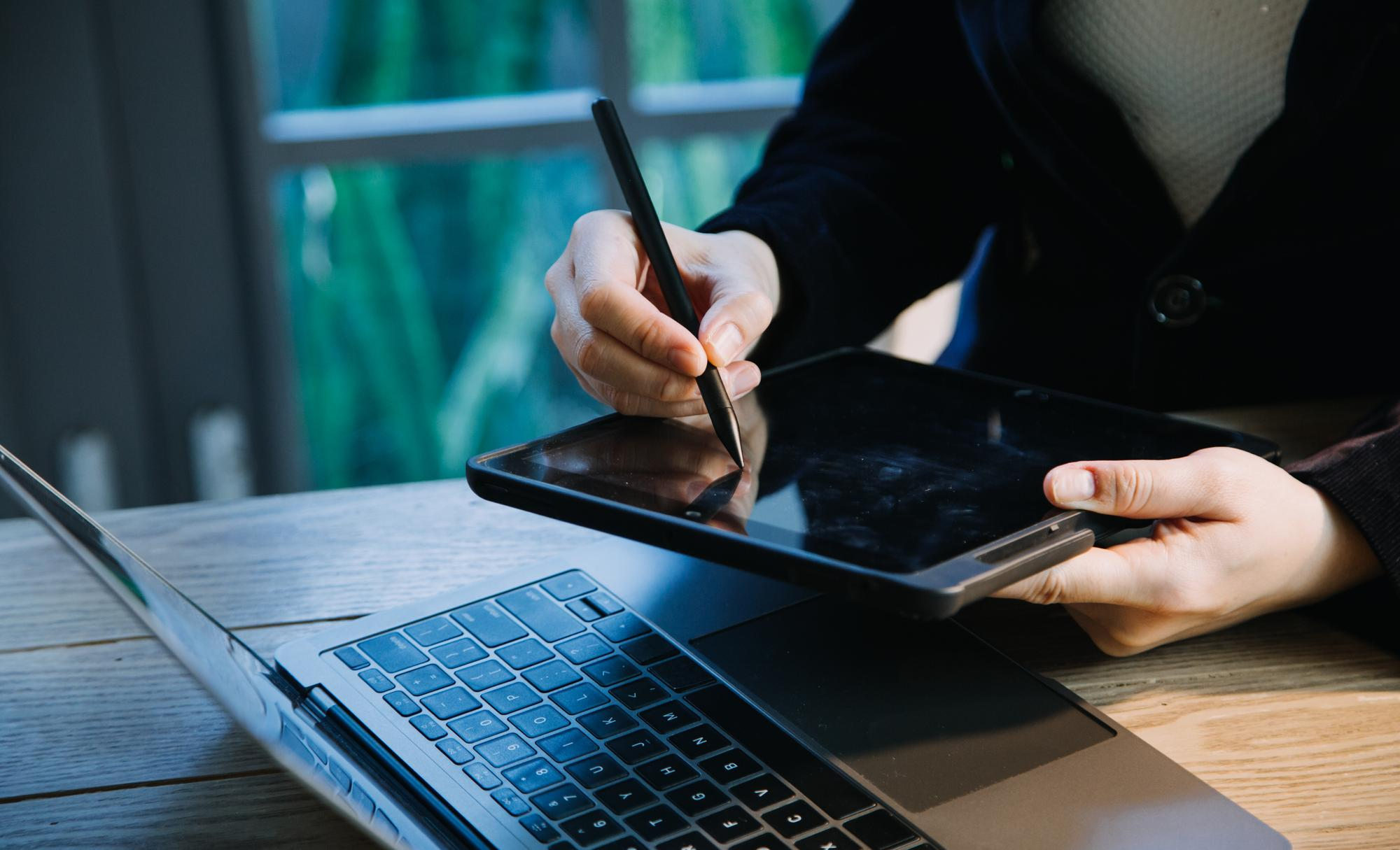 Professional using digital pen on graphics tablet next to laptop on wooden desk for digital design or e-signature work