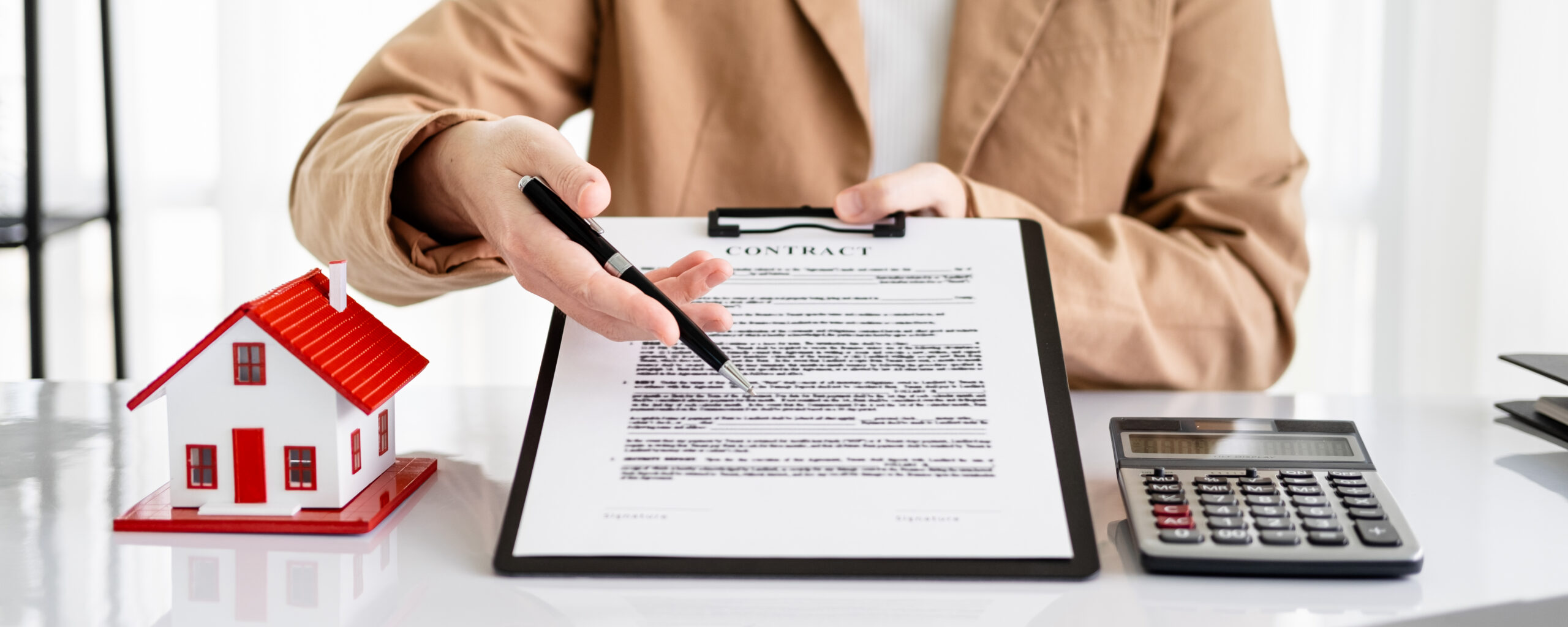 Real estate agent in tan blazer presenting a property contract on clipboard with pen, alongside miniature house model with red roof and calculator on white desk
