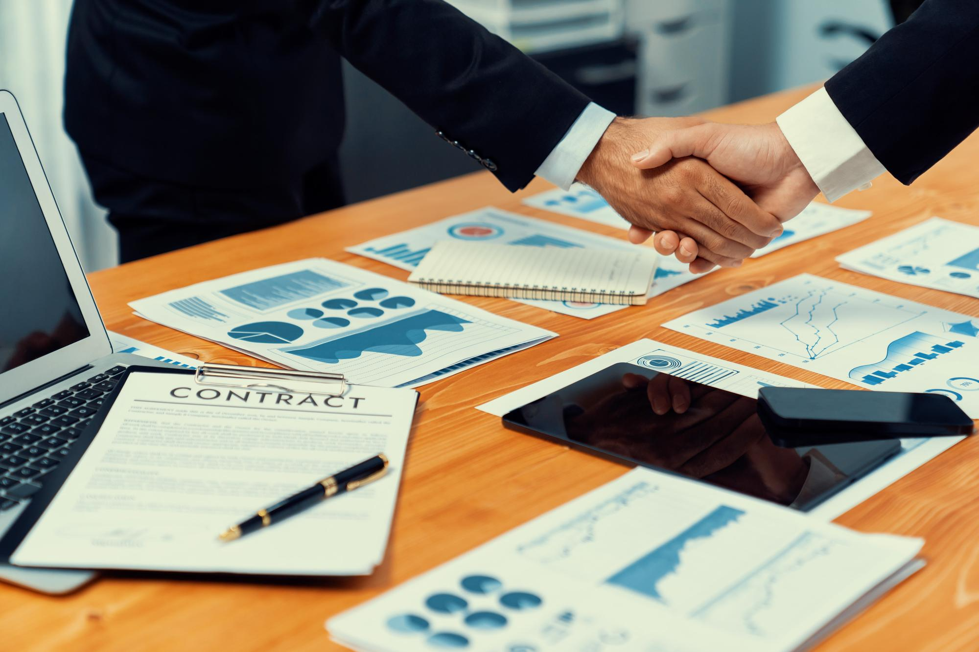 Two business professionals in suits shaking hands over desk covered with contract document, business charts, graphs, laptop, tablet, and pen, sealing a business deal