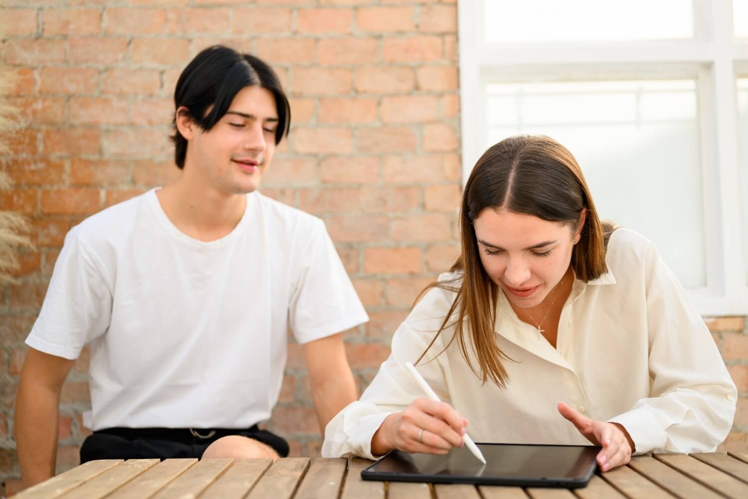 Two people collaborating at a wooden slatted table in a modern space with exposed brick walls - a man in a white t-shirt watches as a woman in a cream blouse uses a white stylus to write on a tablet device